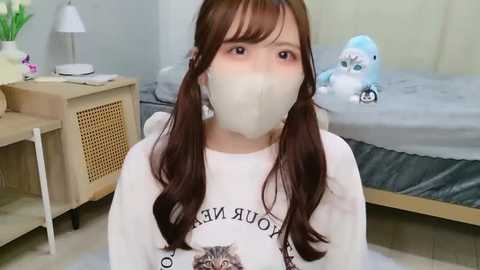 A video of a young Asian woman with long brown hair, wearing a white mask and a graphic tee, sitting on a bed in a cozy, minimalistic bedroom with a stuffed animal and a desk.