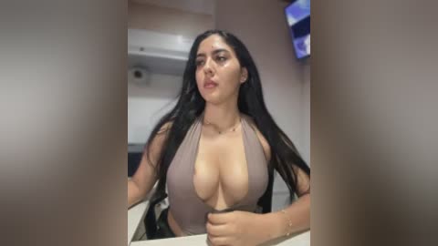 Video of a curvaceous woman with long black hair, wearing a low-cut gray top, seated at a table in a dimly lit room with blurred edges.