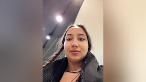 A video of a young woman with medium brown skin, long dark hair in braids, wearing a black jacket and necklace, smiling slightly, indoors with blurred background.