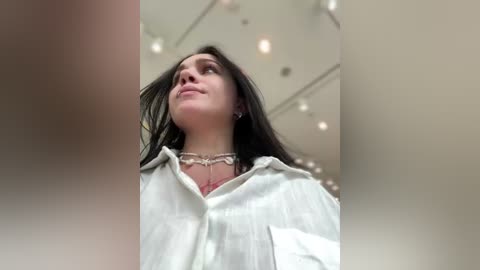 Video of a young woman with long dark hair, wearing a white shirt and necklace, looking up in a modern, well-lit room with overhead lights and soft focus.
