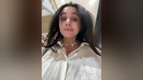 Video of a young woman with long, dark hair, wearing a white shirt and a necklace, standing indoors with a blurred background.