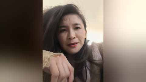 A video of a distraught Asian woman with long dark hair, wearing a beige cardigan, pointing at the camera, set against a blurred, beige background.