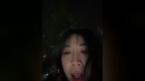 A dimly-lit, grainy video of a young Asian woman with long black hair and fair skin, wearing a dark, possibly wet, garment, appears distressed with a grim expression, mouth agape, eyes wide open.