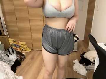 A video of a plus-sized woman in a light gray bra and shorts standing in a messy bedroom with wooden furniture, clothes scattered on the floor.