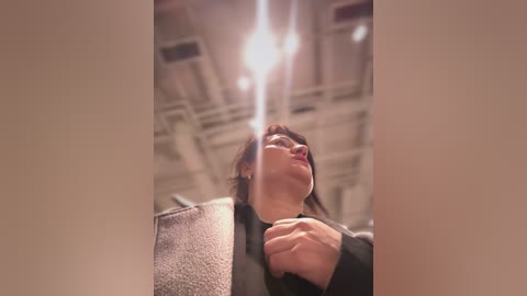 Video of a person with light skin and straight, dark hair, wearing a black sweater, gazing upward. The background features a ceiling with exposed ductwork and fluorescent lights, suggesting an industrial or utilitarian setting.