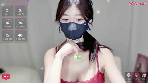 Video of a young Asian woman with fair skin, wearing a red lace bra, black face mask, and black hair in a ponytail, standing in a softly lit room.
