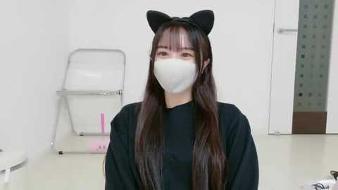 Video of an Asian woman with long, dark hair, wearing a black headband with cat ears, a black robe, and a white surgical mask, in a clinical setting with a white wall, shelves, and a mirror.