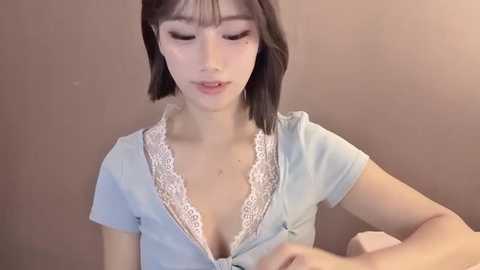 Video of a young Asian woman with straight black hair and fair skin, wearing a light blue shirt with lace trim, unbuttoned to reveal cleavage, against a beige wall.