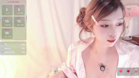 Video of a young Asian woman with fair skin, light brown hair, wearing a pink satin robe, with a visible pubic hair patch and a hair clip, in a soft-lit, minimalist room.