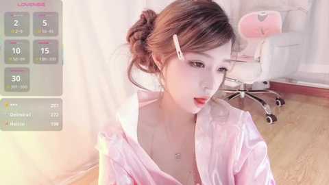 A young Asian woman with fair skin, wearing a pink satin robe, sits in a modern office with a white chair, wooden floor, and light-filled room.