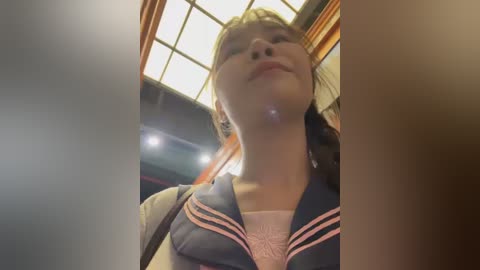 Video of a young Asian woman with light skin, wearing a blue sailor collar shirt, smiling, taken from a low angle, showing off her neck and chest, inside a dimly lit room with wooden beams and a grid ceiling.