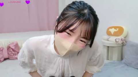 A video of an Asian woman with dark hair, wearing a white dress and a face mask, kneeling on a bed, blushing, in a pastel-colored room.