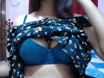 Video of a woman with medium skin tone, wearing a blue bra with a floral pattern, lifting a floral shirt, revealing cleavage. Background shows a bed with blue and white sheets, a wooden door, and red curtains.