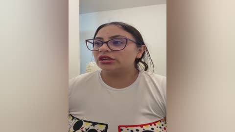 Video of a Latina woman with medium skin tone, brown hair, wearing glasses, a white t-shirt with a graphic design, and a neutral expression.