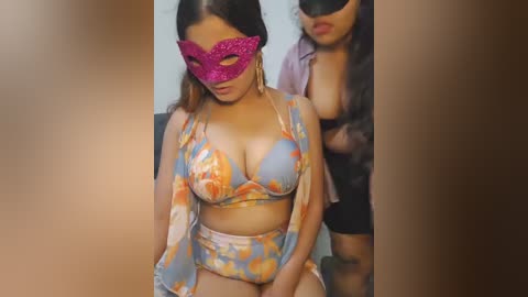 Video of a Latina woman with medium skin tone, wearing a colorful floral bikini, purple masquerade mask, and sheer sarong, sitting indoors.