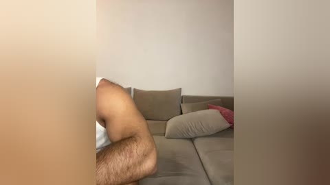 A video of a man with a hairy, muscular arm resting on a beige couch in a minimalist room with neutral-toned walls.