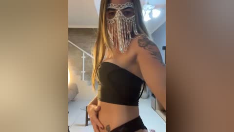 Video of a light-skinned woman with long blonde hair, wearing a black strapless crop top, a beaded face mask, and black shorts, with a tattoo on her left shoulder, indoors against a beige wall.