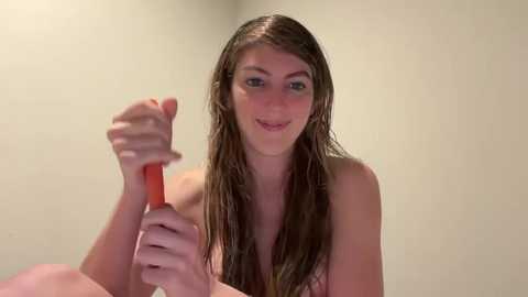 Video of a young Caucasian woman with long, wet brown hair, holding a red comb in her right hand, smiling. She is topless, with a beige wall background.