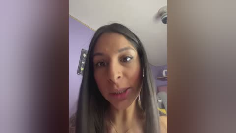 Video of a young woman with medium-brown skin and straight, long black hair, wearing a pink tank top. She has a slightly open mouth and looks surprised. The background shows a purple wall with a framed photo and a ceiling light.