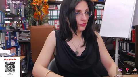 Video of a fair-skinned woman with shoulder-length black hair, wearing glasses, a black blouse, and a necklace, seated in a library with bookshelves and flowers in the background.