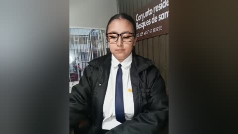 Video of a Latina woman with dark hair in a bun, wearing glasses and a black jacket over a white shirt and dark tie, standing in front of a sign with Spanish text, in a modern office setting.