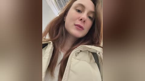 A video of a fair-skinned woman with long auburn hair, wearing a beige puffer jacket, standing indoors with a blurred background. She has a neutral expression and is looking slightly to the side.