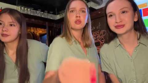 Video of three women in light green shirts, with one holding a pink drink, in a cozy, dimly lit bar. Background features dark wooden walls and colorful chalkboard art.