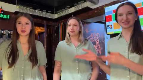 Video of three young women, two with long hair, in light green tops, standing in a cozy bar with wooden walls, colorful graffiti, and a TV screen in the background.