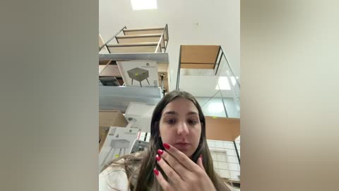 Video of a young woman with long brown hair, fair skin, and red-painted nails, standing in a modern, partially constructed building with wooden scaffolding, glass railings, and white walls.