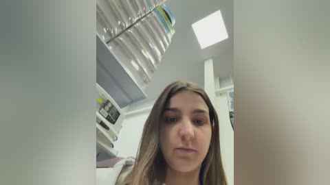 A video of a young woman with light skin and long, straight brown hair, looking serious, standing in a modern, well-lit kitchen with white cabinets, a glass shelf, and a large window.
