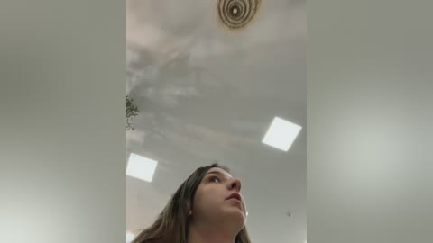 A video of a young woman with long brown hair, looking up, in a room with a circular ceiling light and white walls.