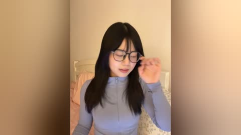 Video of an Asian woman with long black hair and glasses, wearing a light blue zip-up sweater, adjusting her glasses in a dimly lit bedroom with beige walls and a bed with floral-patterned sheets.