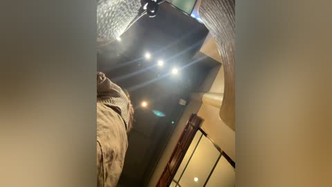 Video of a person in a beige coat, partially obscured by a textured, beige ceiling light fixture, with a soft, warm indoor light.