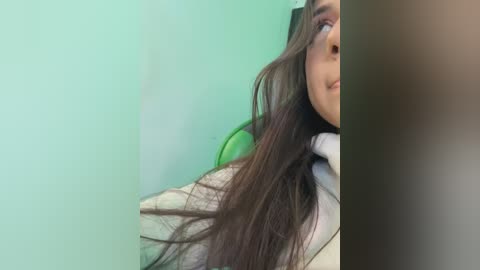 Video of a woman with long, straight brown hair, partially covering her face, wearing a white shirt, sitting against a green wall.