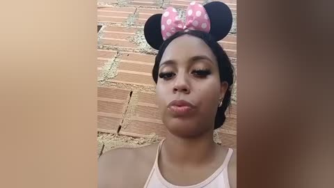 Video of a young woman with medium brown skin, wearing a white tank top, and a Minnie Mouse headband with polka dots, standing against a brick wall.