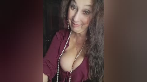 Video of a smiling, light-skinned woman with curly brown hair, wearing a deep burgundy top revealing ample cleavage and a long pearl necklace. The background is blurred, featuring dark colors.