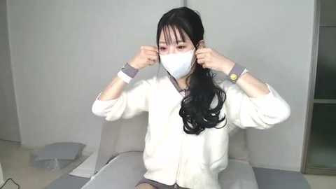 Video of an East Asian woman with long black hair, wearing a white mask, adjusting it with both hands. She is in a sterile, white room with a table and a chair.