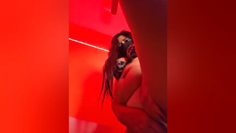 Video of a naked woman with long hair, bound with tape, standing in a red-lit room. Her face is obscured by a black mask. The image conveys a sense of vulnerability and restraint.