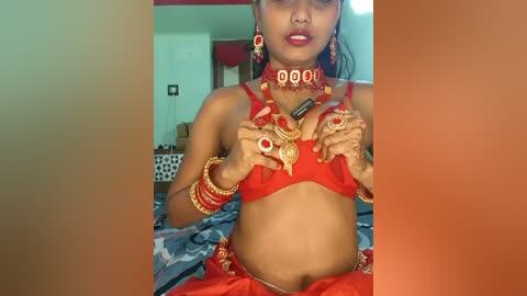 Video of a young South Asian woman in a red traditional outfit, adorned with gold jewelry, including a bindi, earrings, and a necklace, in a bedroom with a bed and red curtains.