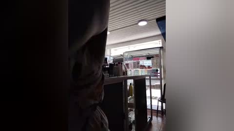 Video of a dimly-lit interior, featuring a person's shadowy figure on the left, with shelves of items in the background, and a brightly lit storefront visible through a glass door on the right.