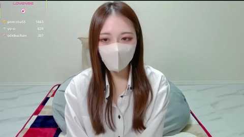 A video of an East Asian woman with long brown hair, wearing a white surgical mask and white top, kneeling on a bed with a colorful quilt, against a plain white wall.