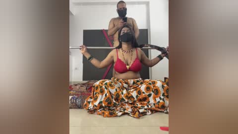 Video of a Black woman with medium skin tone, wearing a red bra and colorful skirt, being held by a muscular Black man with a shaved head, in a modern living room.