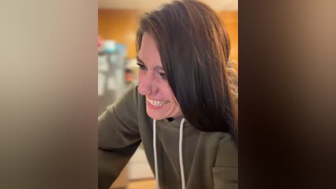 Video of a middle-aged woman with long dark hair, smiling while using a computer, wearing a green hoodie, in a warmly lit, blurred indoor setting.