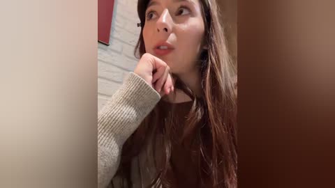 Video of a young woman with long brown hair, wearing a beige sweater, leaning against a wall, gazing introspectively.