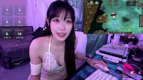 Video of an Asian woman with long black hair, wearing a white lace top, seated at a cluttered desk with gaming equipment and a TV displaying a game.