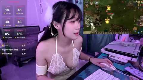 A video shows an Asian woman with long black hair and bangs, wearing a revealing white lace top, playing a video game on a computer in a dimly lit room.