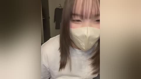 A video of a person with long, straight brown hair wearing a white long-sleeve shirt and a yellow face mask, standing in a dimly lit hallway with beige walls and a dark coat hanging on a hook.