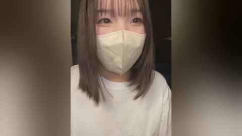 Video of an Asian woman with straight, shoulder-length brown hair, wearing a white t-shirt and a beige surgical mask. Background is blurred with a dark tone.