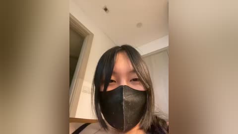 Video of an Asian woman with straight, shoulder-length black hair, wearing a black face mask and dark top, standing in a well-lit room with white walls and a door.