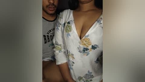 Video of a man with a trimmed beard, wearing a \"Brooklyn\" t-shirt, and a woman with medium brown skin, wearing a floral-patterned white nightgown, seated closely together.
